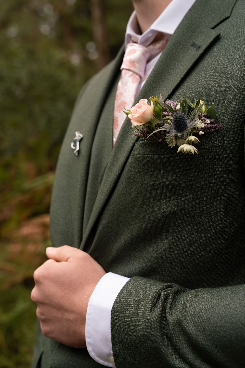 Close-up details of a dark green suit jacket with rustic floral boutonniere against a natural background.