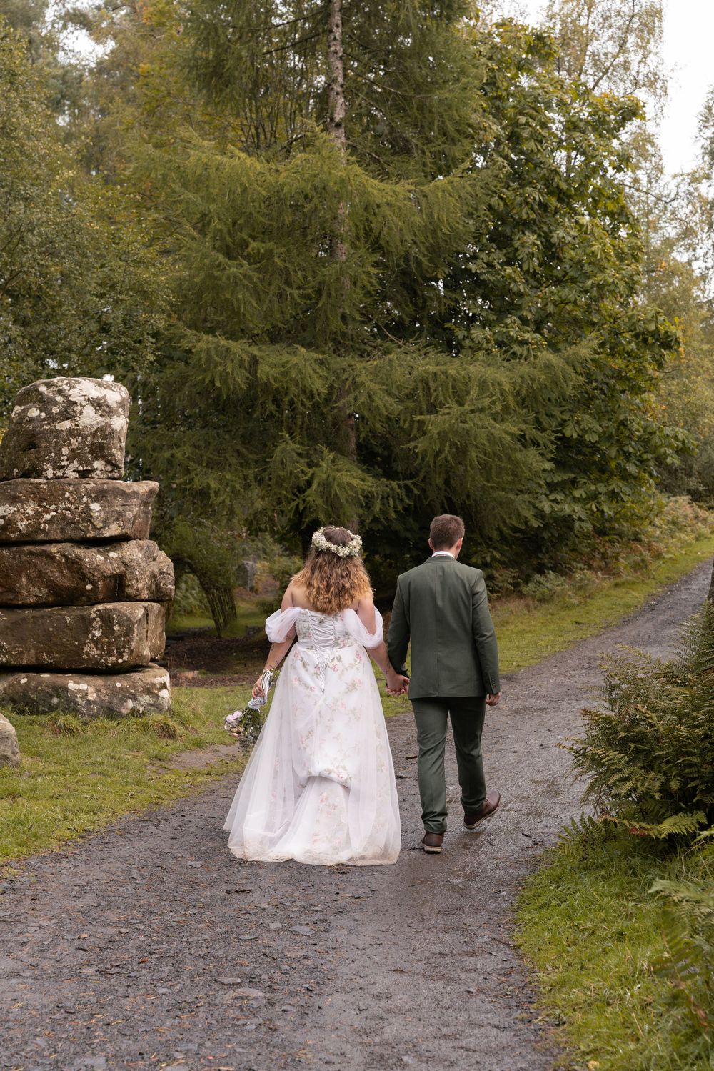 A couple in wedding attire walking down a gravel path surrounded by lush greenery and stone pillars.