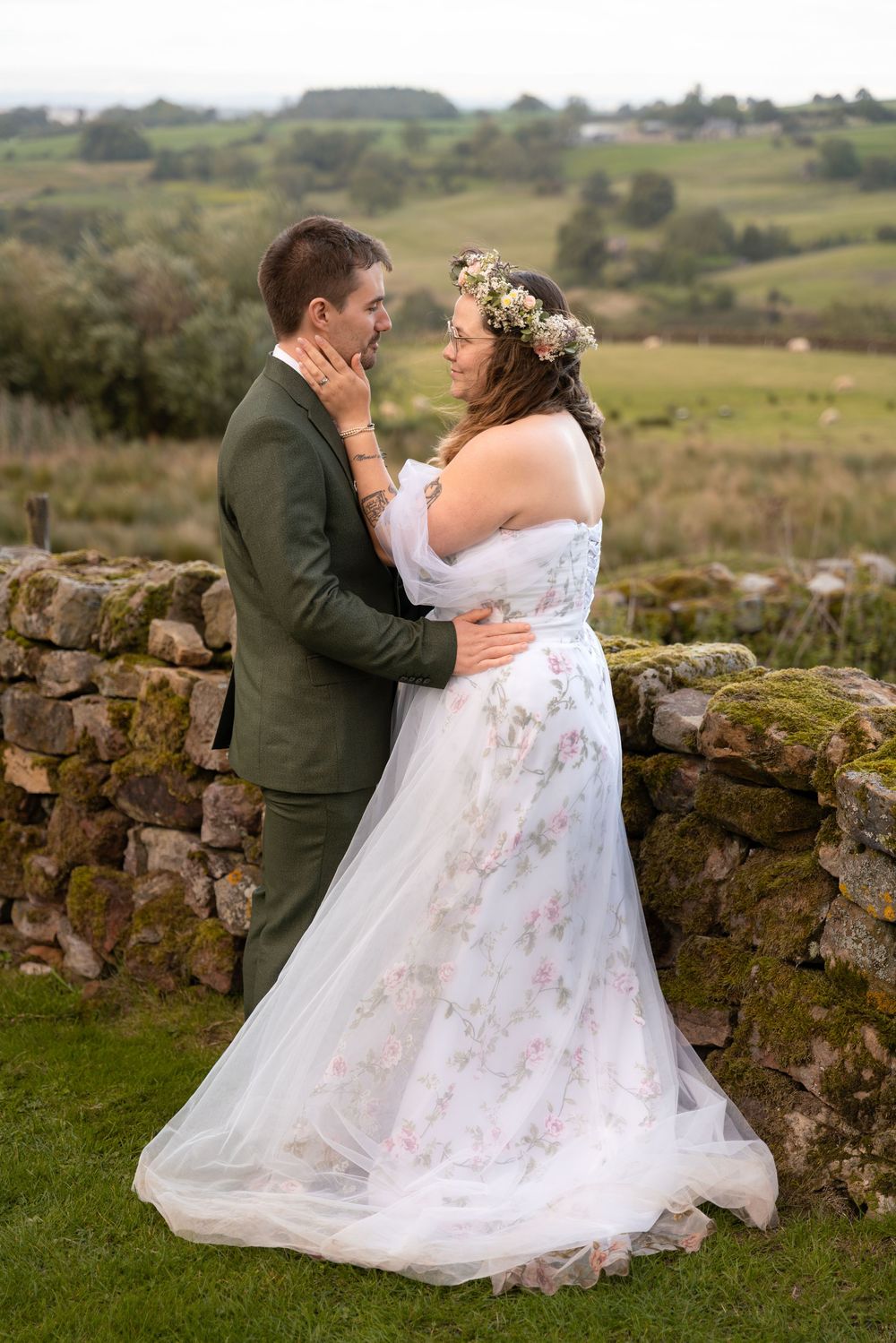 Romantic wedding couple embraces by rustic stone wall in countryside setting during outdoor photoshoot.