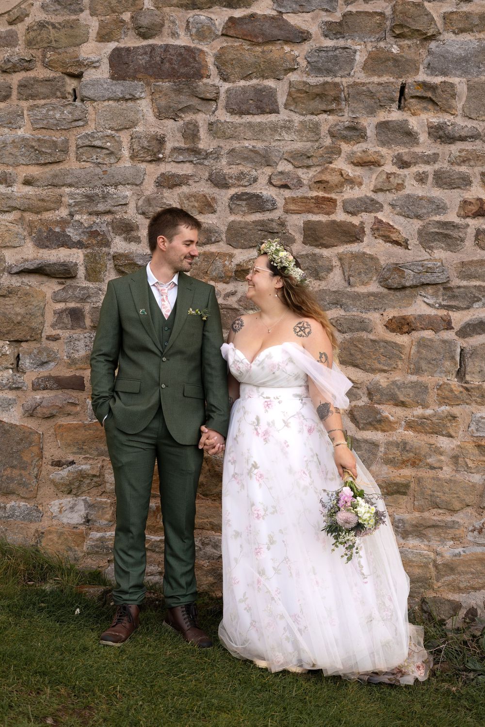 Wedding couple poses against rustic stone wall backdrop in elegant formal attire.