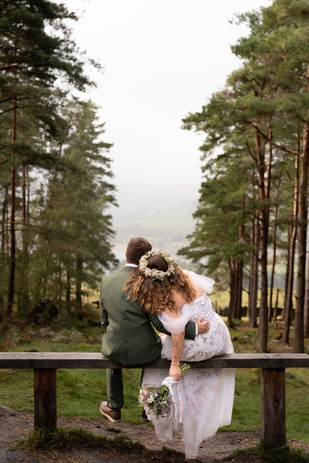 A romantic couple shares intimate moments on a wooden bench surrounded by tall pine trees in a misty forest setting.