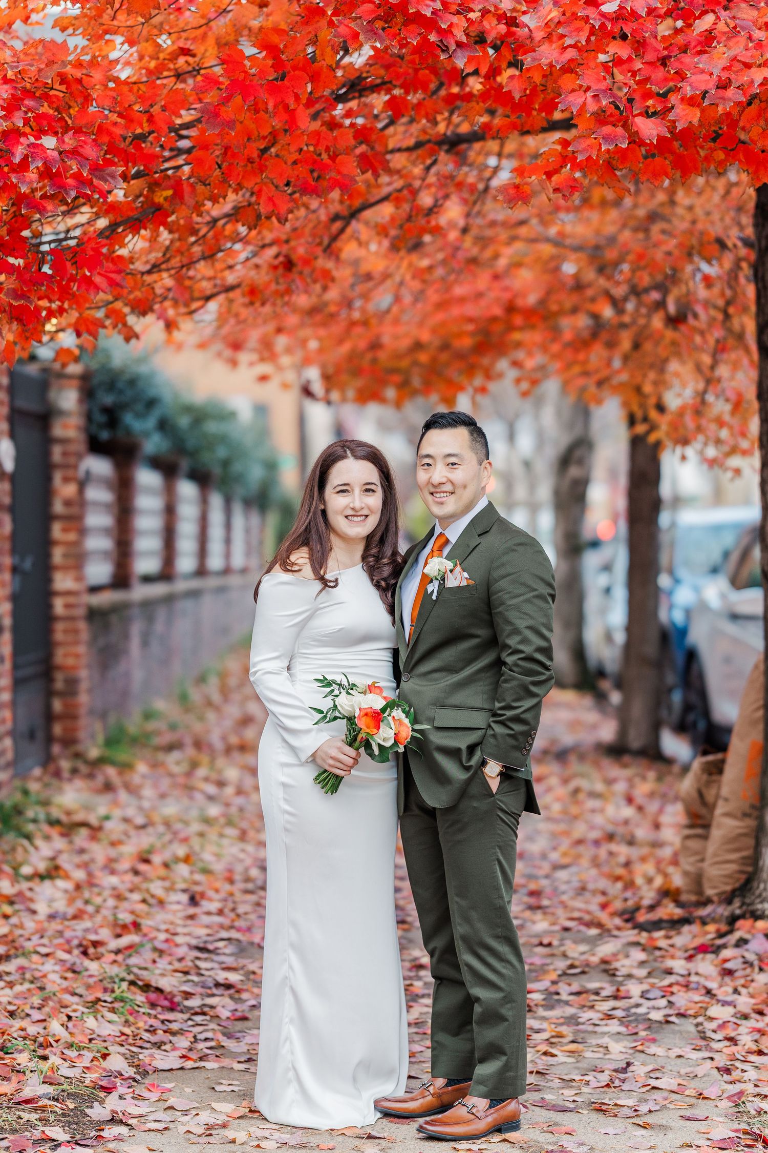 A couple poses for wedding photos under vibrant red autumn trees with fallen leaves scattered on the ground.