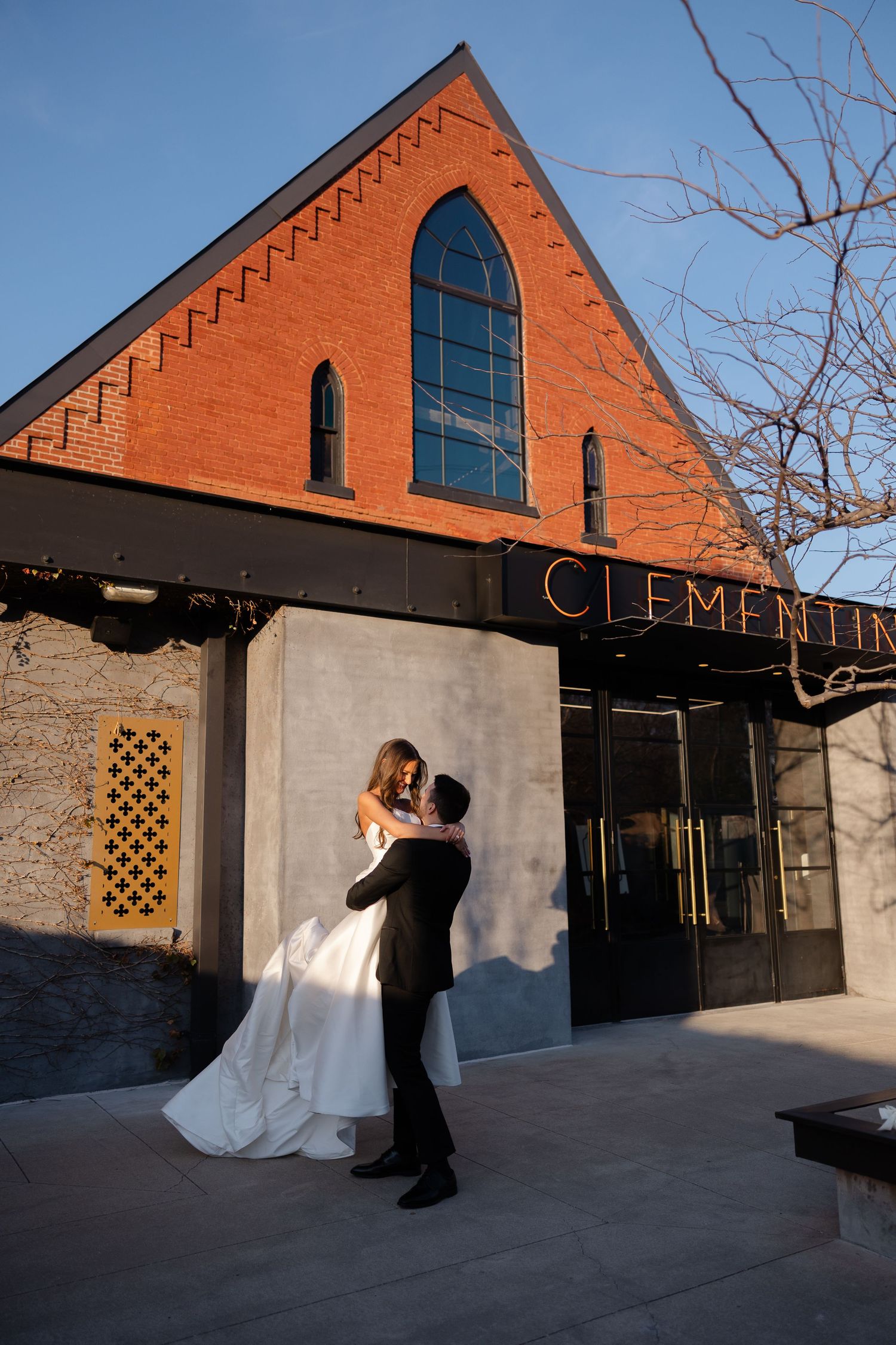 A couple in formal wedding attire poses in front of a rustic brick building with arched windows at sunset.