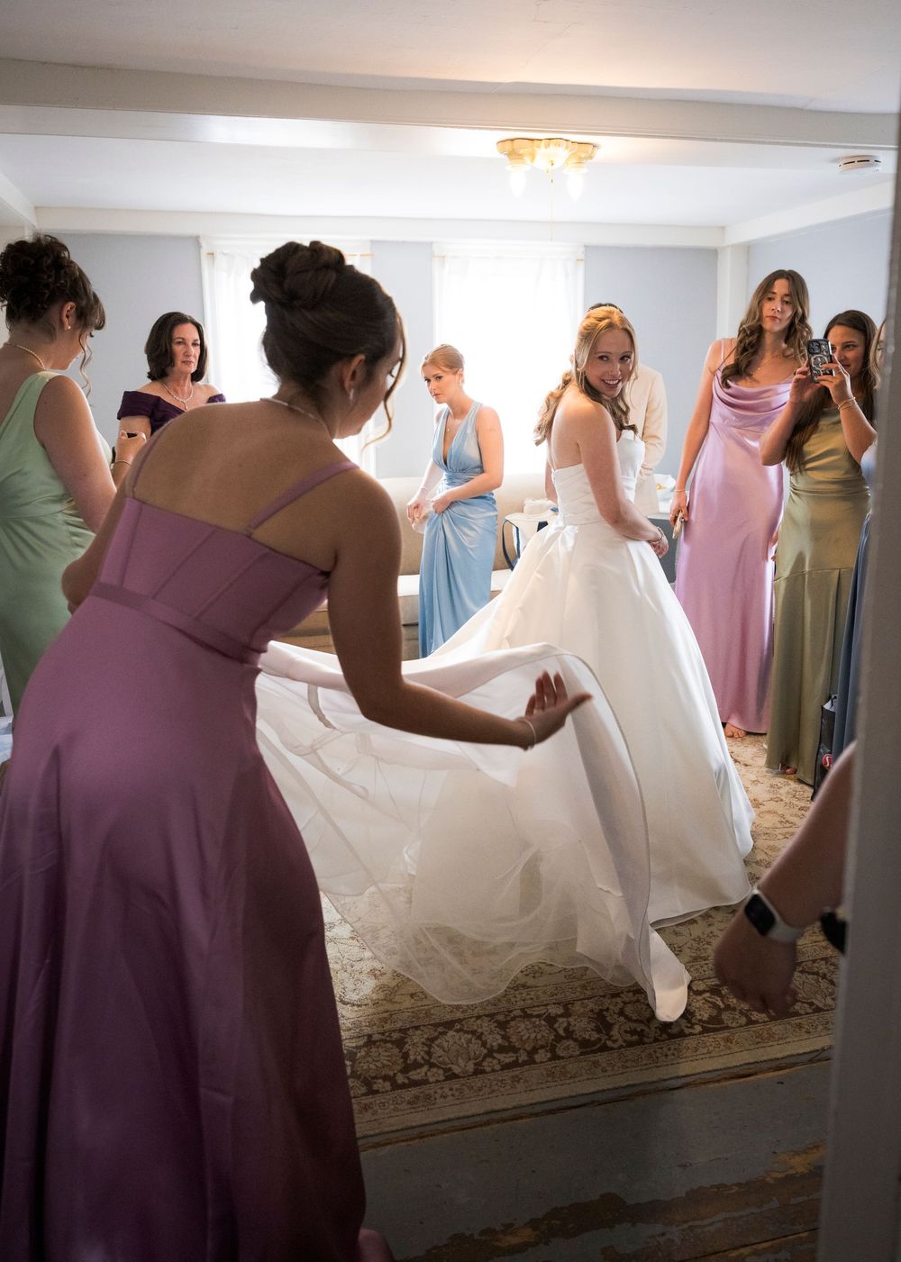 Bridesmaids in pastel gowns help bride with wedding dress during bridal preparations.