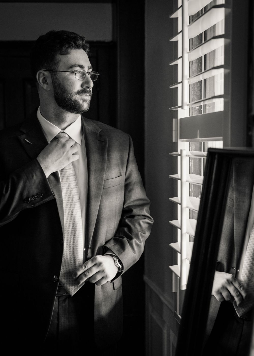 Groom adjusts tie while looking out window in black and white getting ready photo.
