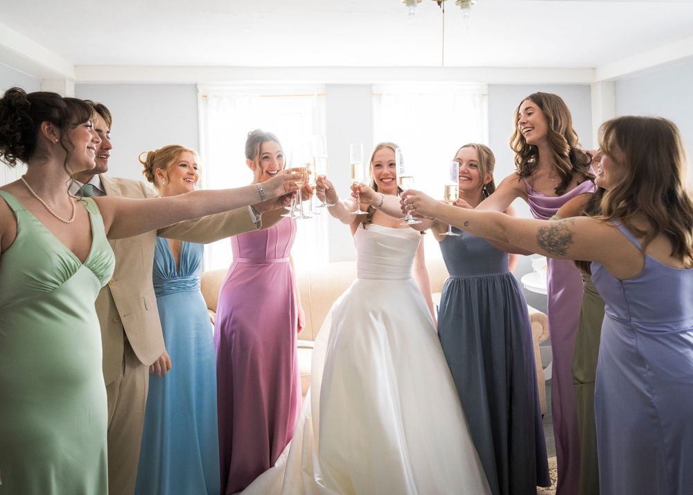 Bride in white gown toasts with bridesmaids in colorful pastel dresses during pre-wedding celebration indoors.