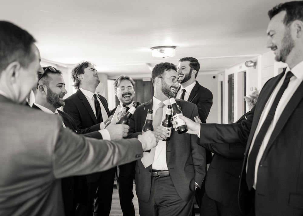 Groom celebrates with groomsmen toasting drinks in black and white wedding photo.