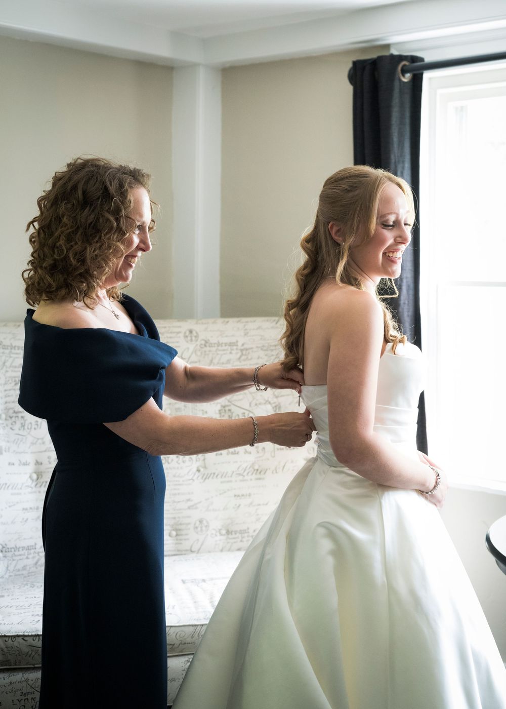 Mother helps bride button her white wedding gown in a bright, elegantly decorated dressing room.