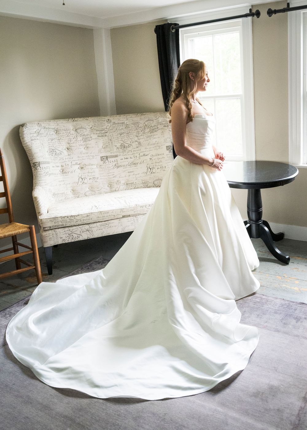 Bride in elegant white ball gown with long train standing by window in bridal suite.