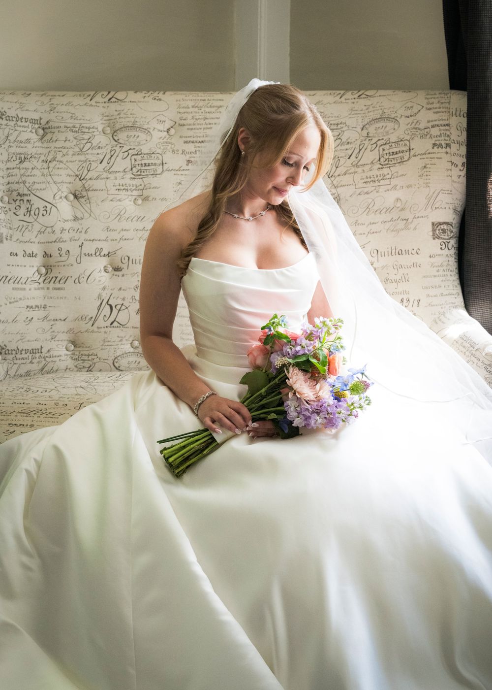 Elegant bride in a strapless white gown holding a colorful bouquet while seated on a decorative bench.