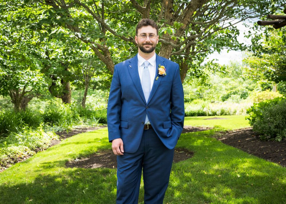 Groom in navy blue suit with yellow boutonniere standing in lush green garden setting.