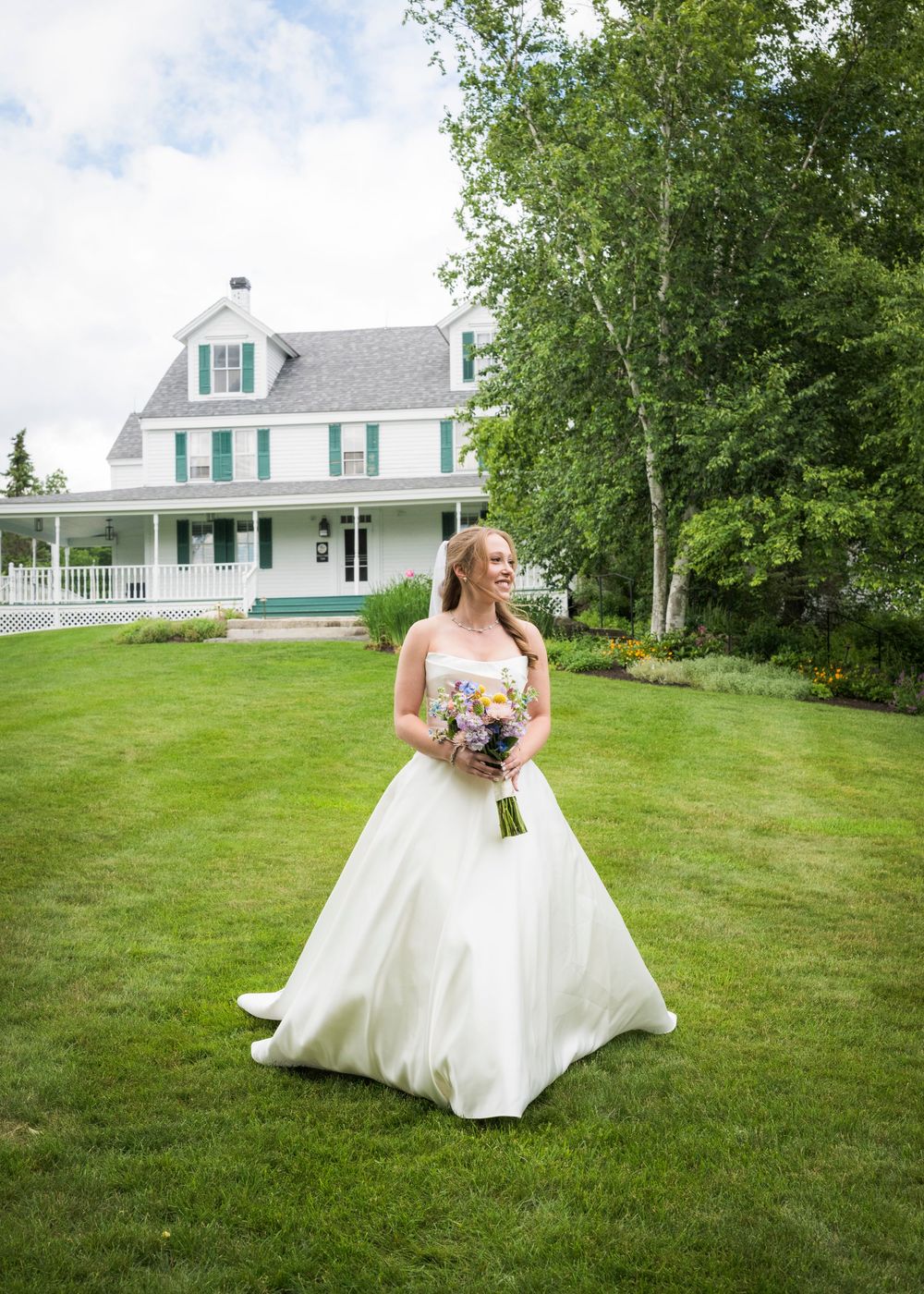 Bride in white ball gown holding bouquet standing on green lawn before white farmhouse venue.