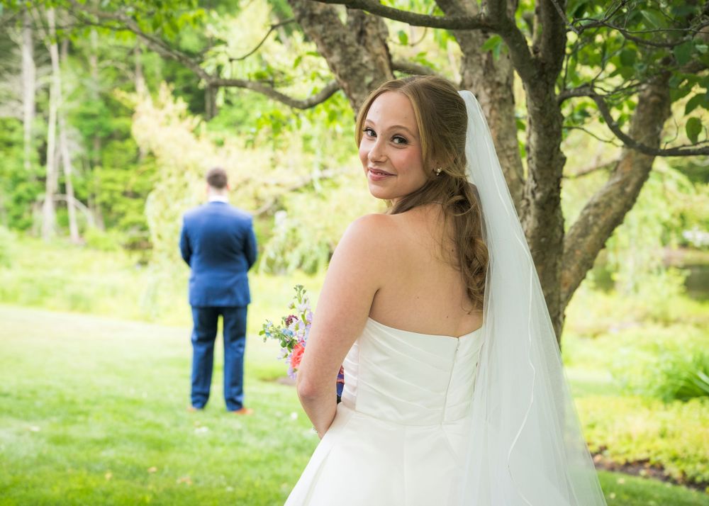 Bride in white gown and veil looks back smiling while groom stands ahead in garden.