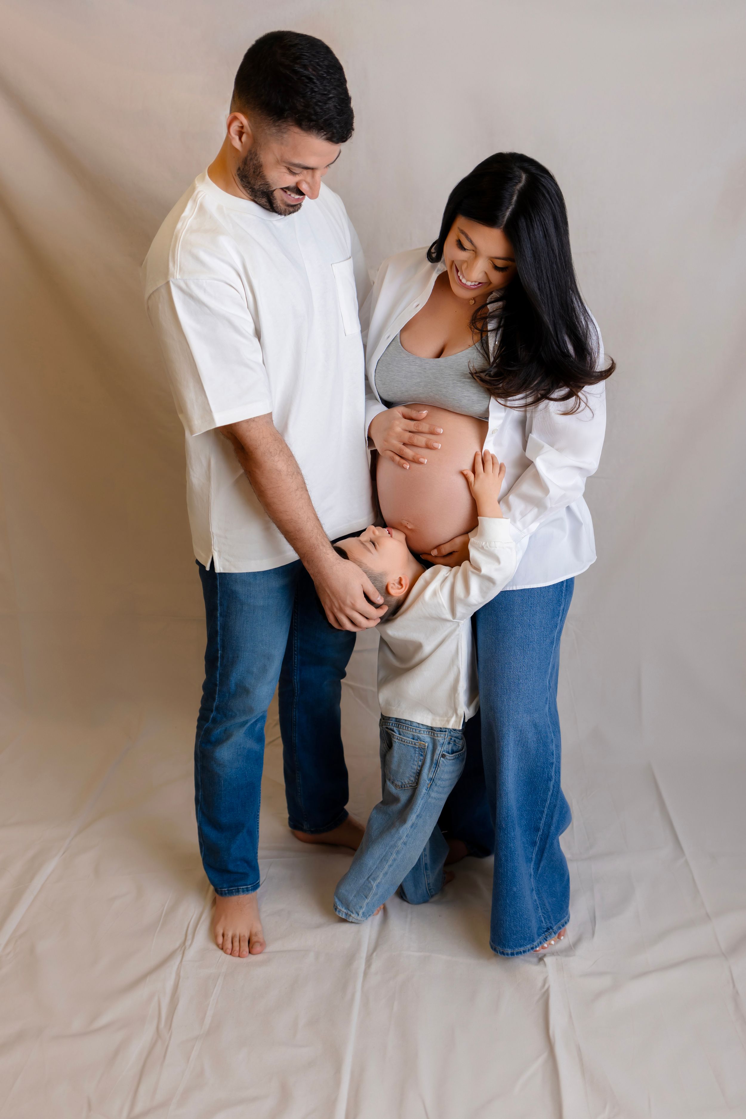 A couple in matching white shirts and jeans embrace while touching a pregnant belly against a neutral backdrop.