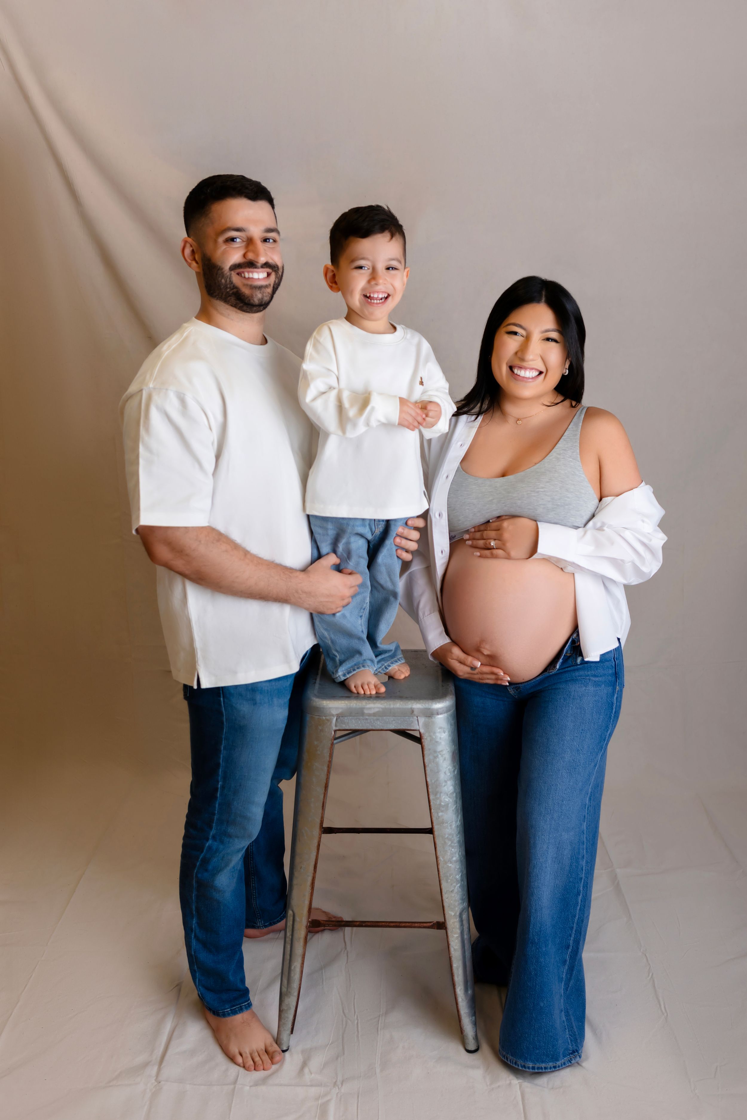 A family of three stands together for a maternity portrait in casual white tops and jeans against a beige backdrop.