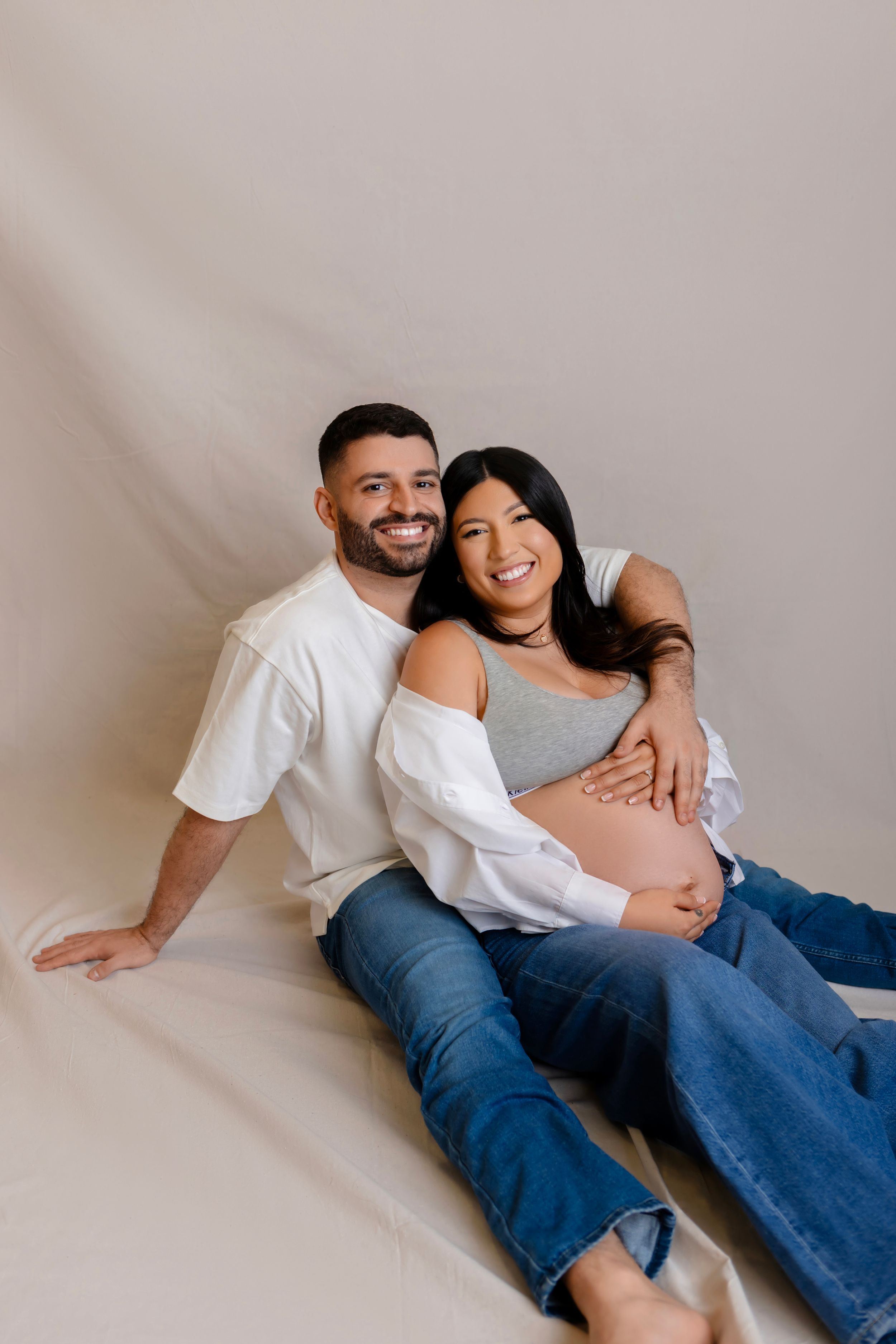 A happy couple in casual attire reclines against a white backdrop during a maternity photoshoot.