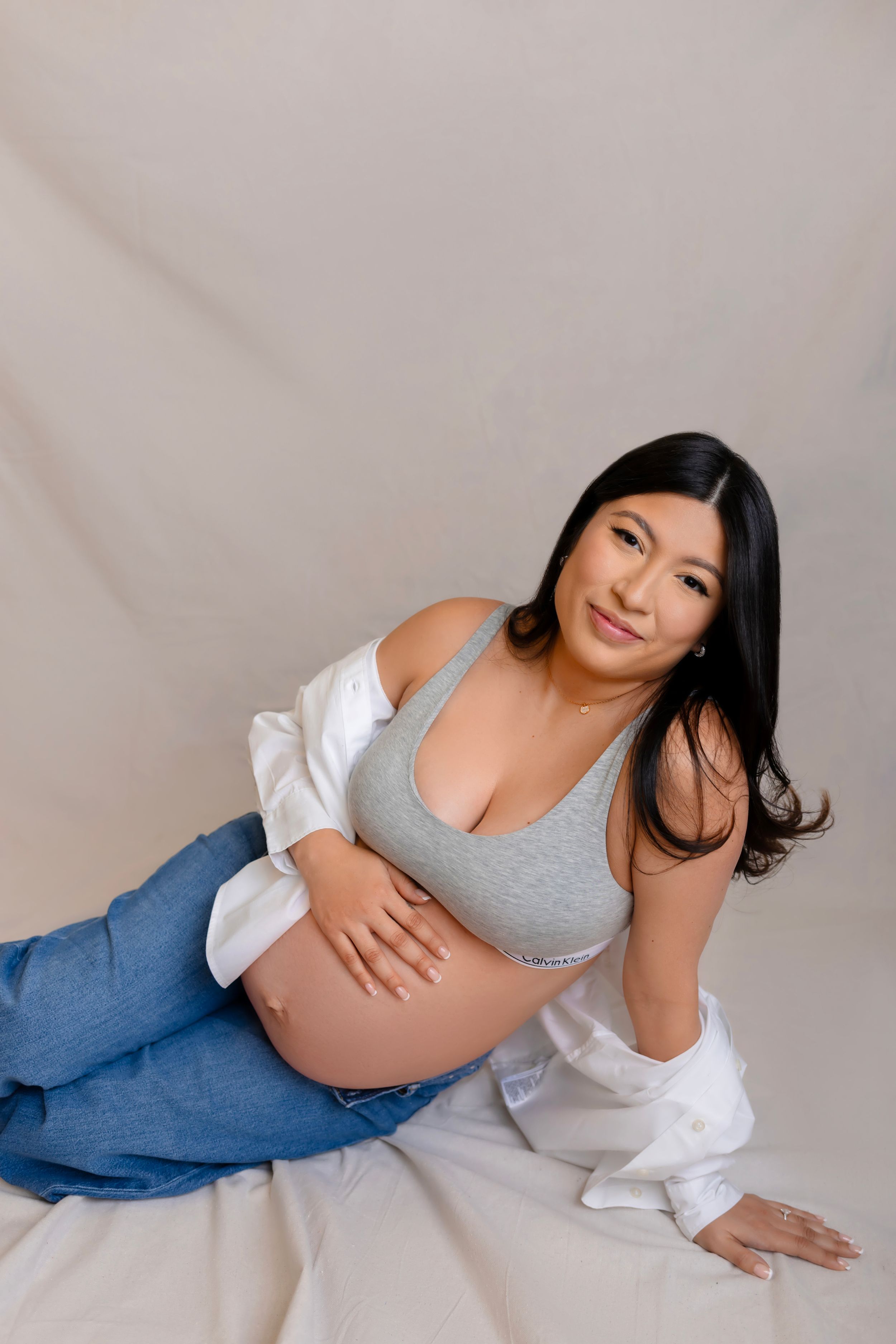 A pregnant woman wearing a gray tank top and blue jeans reclines on a white backdrop during a maternity photoshoot.