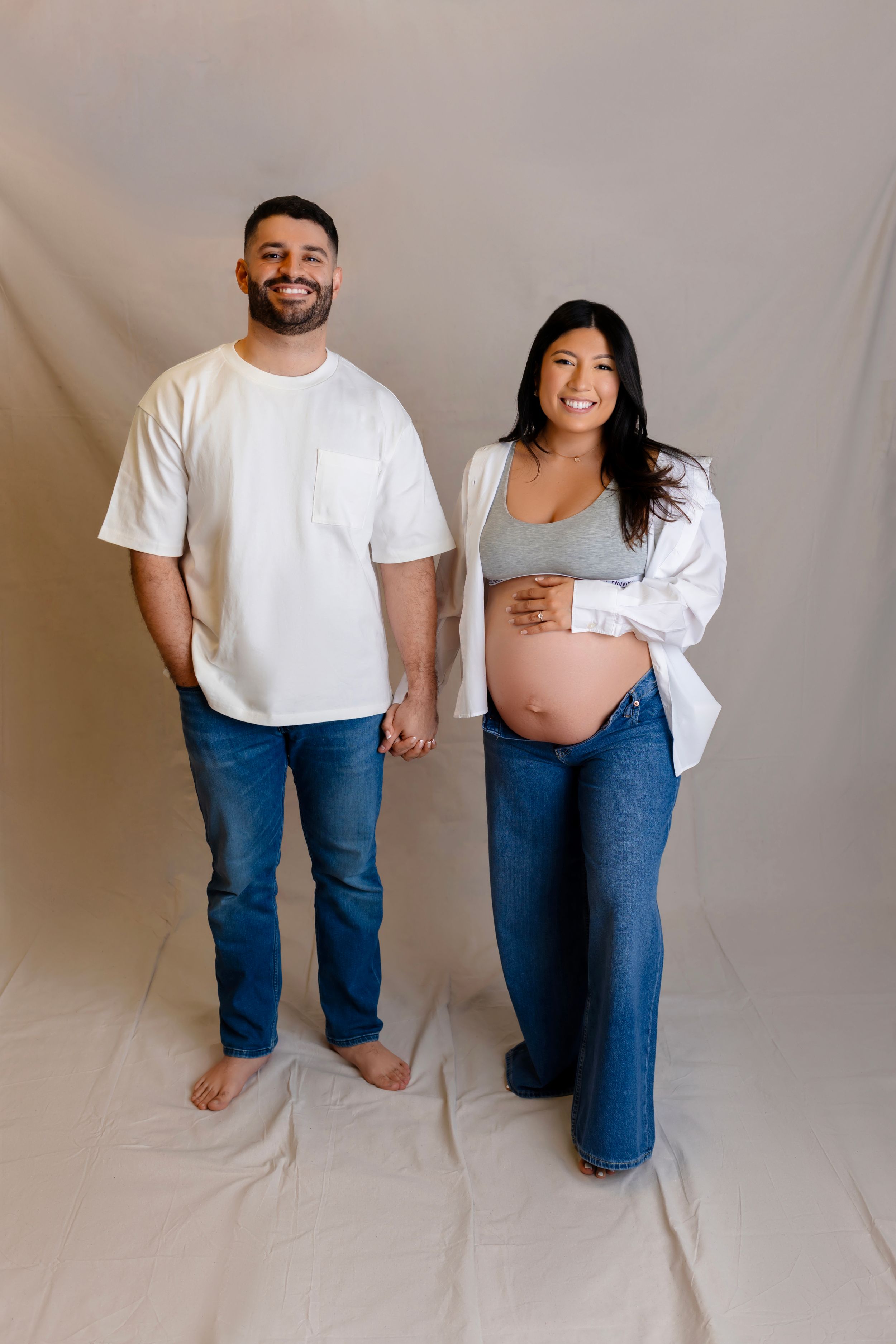 Expectant couple holds hands in casual attire against beige backdrop during maternity photo session.