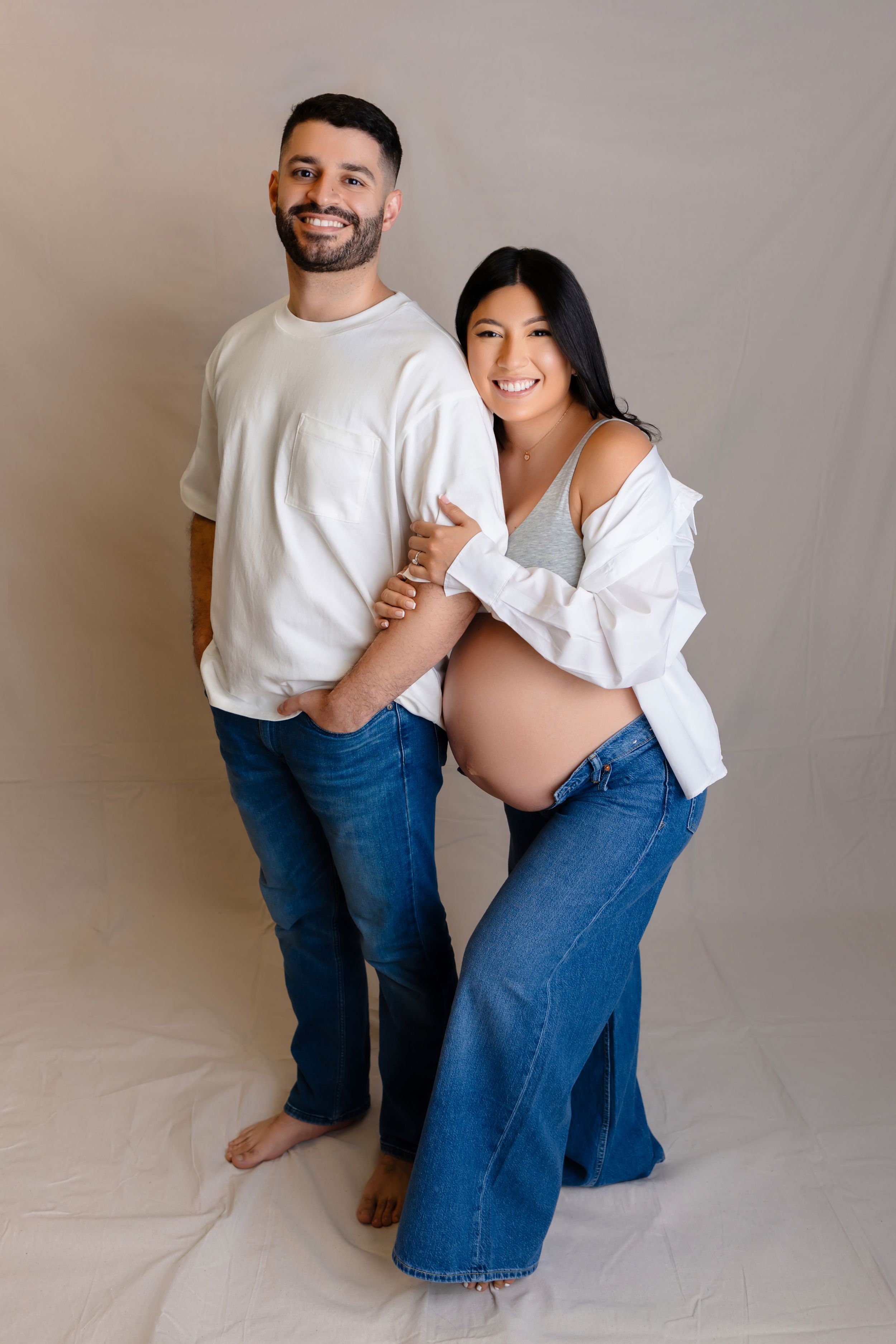 Maternity studio photo of expectant couple wearing casual denim and white tops against neutral backdrop.