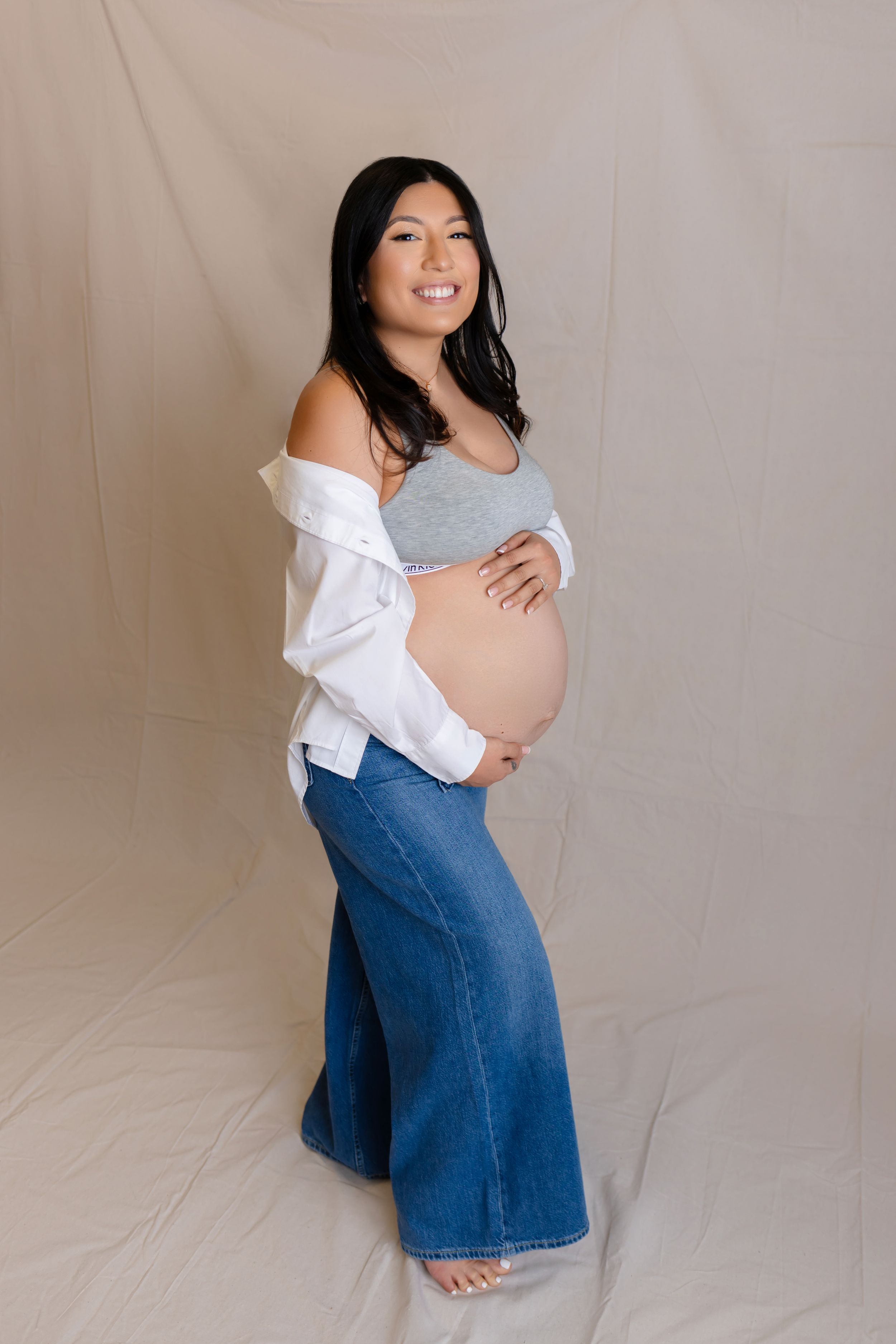 A pregnant woman in blue jeans and gray top poses against a light backdrop during a maternity photoshoot.