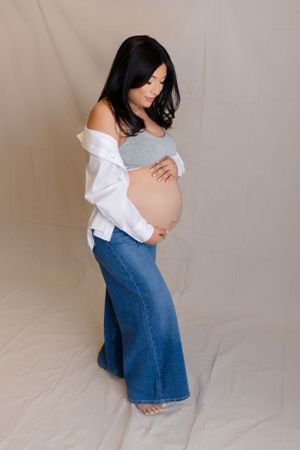 A pregnant woman in a white cardigan, gray top and flared jeans poses sideways against a light backdrop.