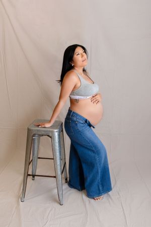 Pregnant woman in casual attire poses beside metal stool against beige backdrop for maternity photo shoot.