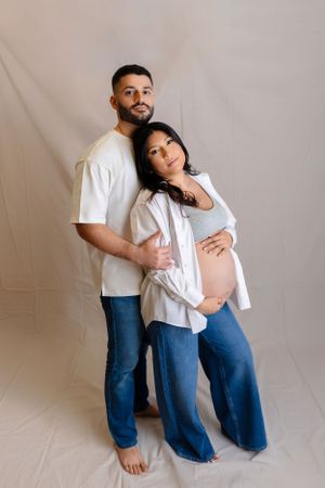 A couple in matching white tops and blue jeans embraces tenderly against a beige backdrop during a pregnancy photoshoot.