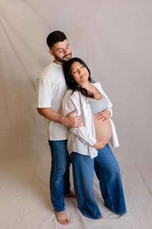 Expecting couple in matching white shirts and jeans embrace lovingly against cream backdrop while celebrating pregnancy.