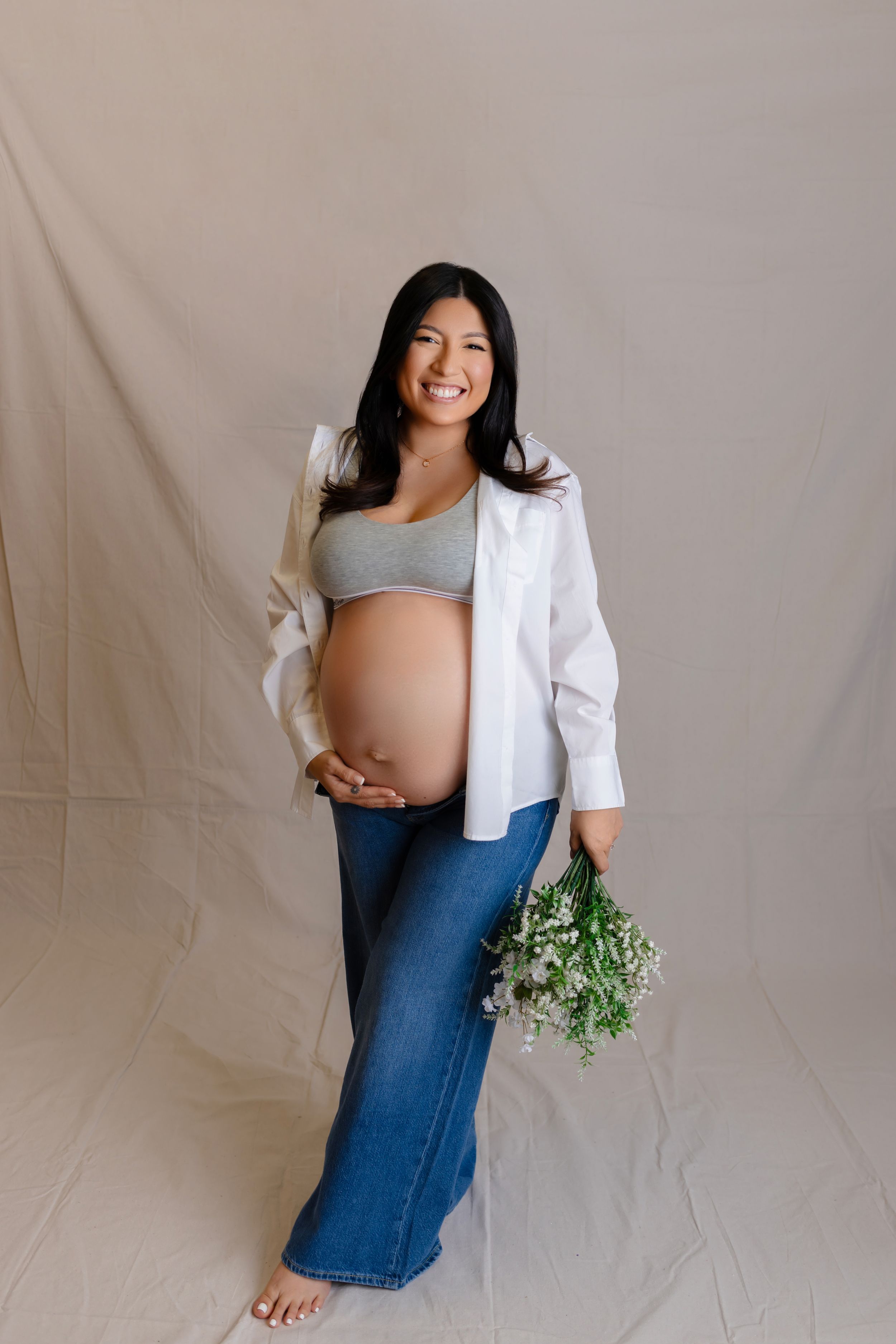 Pregnant woman in white shirt and jeans poses with greenery against neutral backdrop during maternity photoshoot.