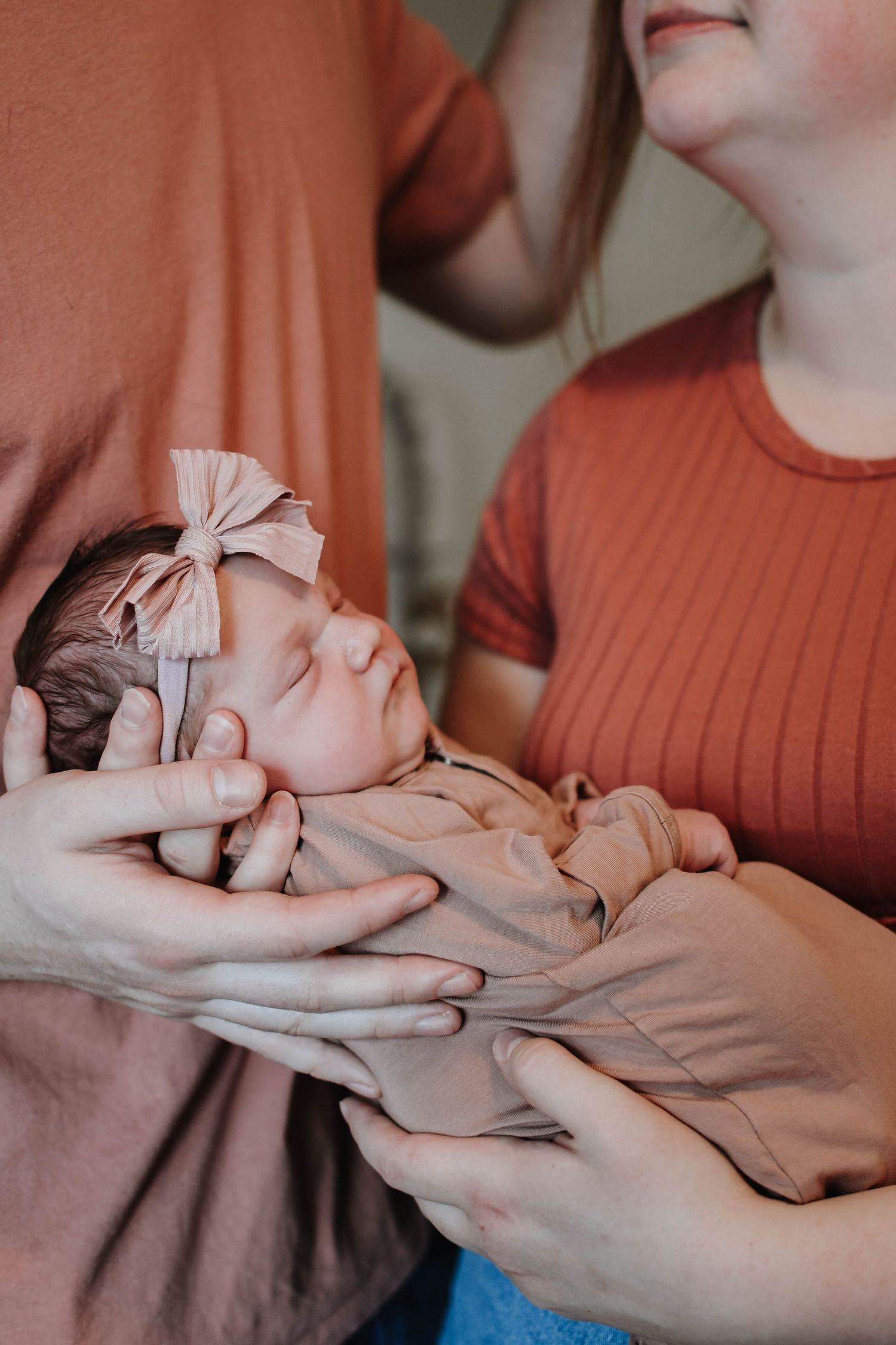 A sleeping newborn baby wearing a pink bow headband is cradled in gentle hands while dressed in neutral tones.