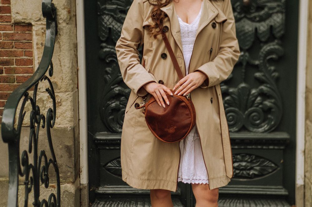 Person in beige coat walking past traditional Dutch buildings with brown leather bag.