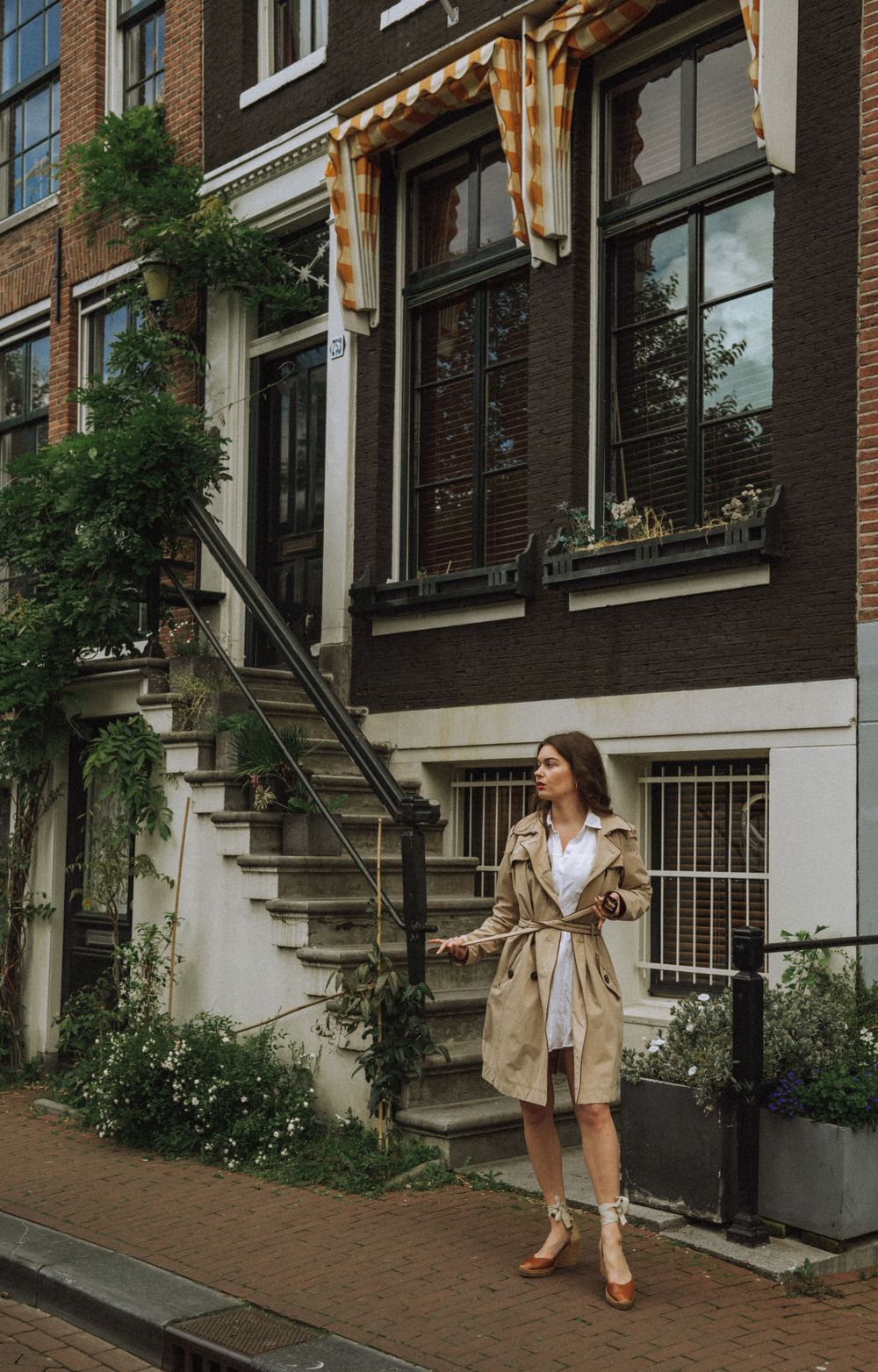 Person in trench coat posing near historic Dutch townhouses with climbing vines.