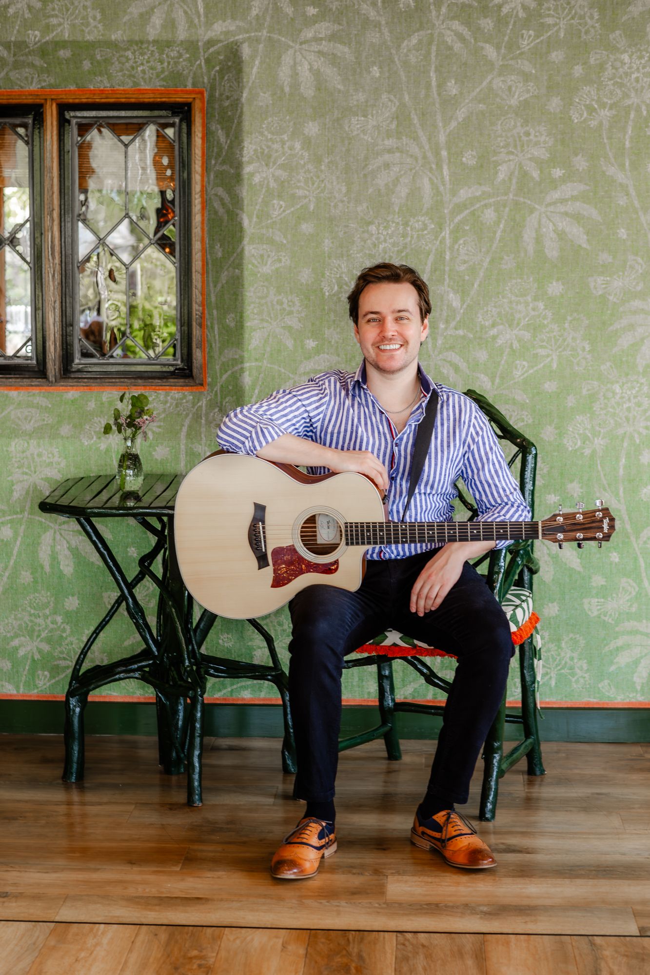 Musician sitting with acoustic guitar against floral wallpaper background.
