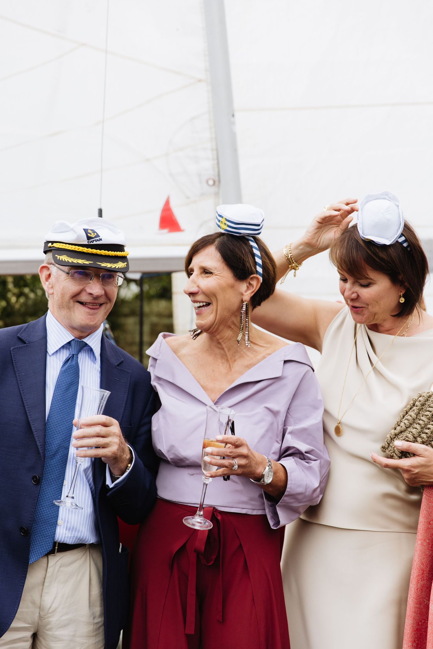Well-dressed guests at an outdoor event share drinks and laughter while wearing nautical-inspired headwear.