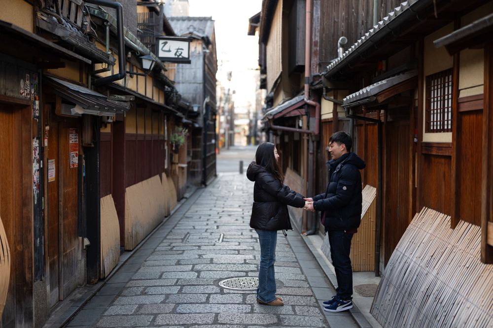 Couple holds hands on a quiet traditional Japanese alleyway surrounded by wooden machiya buildings in Kyoto.