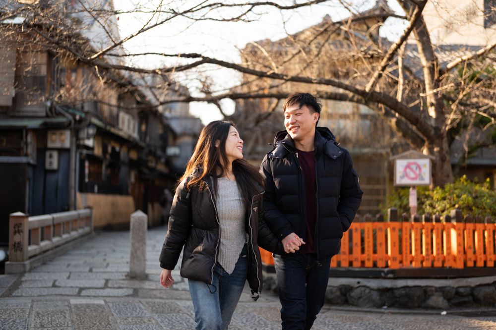 Happy Asian couple walk hand in hand along a scenic Japanese street with bare winter trees and traditional buildings.