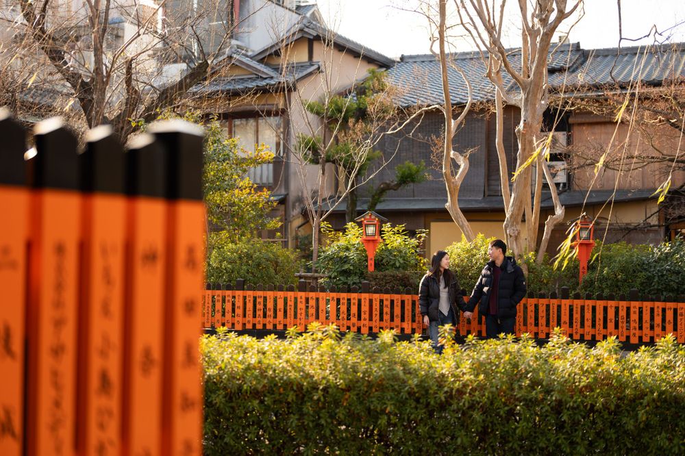 Couple strolls near vibrant orange torii gate pillars and yellow winter flowers at a Japanese shrine garden.