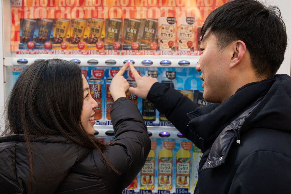 Young Asian couple laugh together while browsing a colorful Japanese vending machine filled with snacks and drinks.