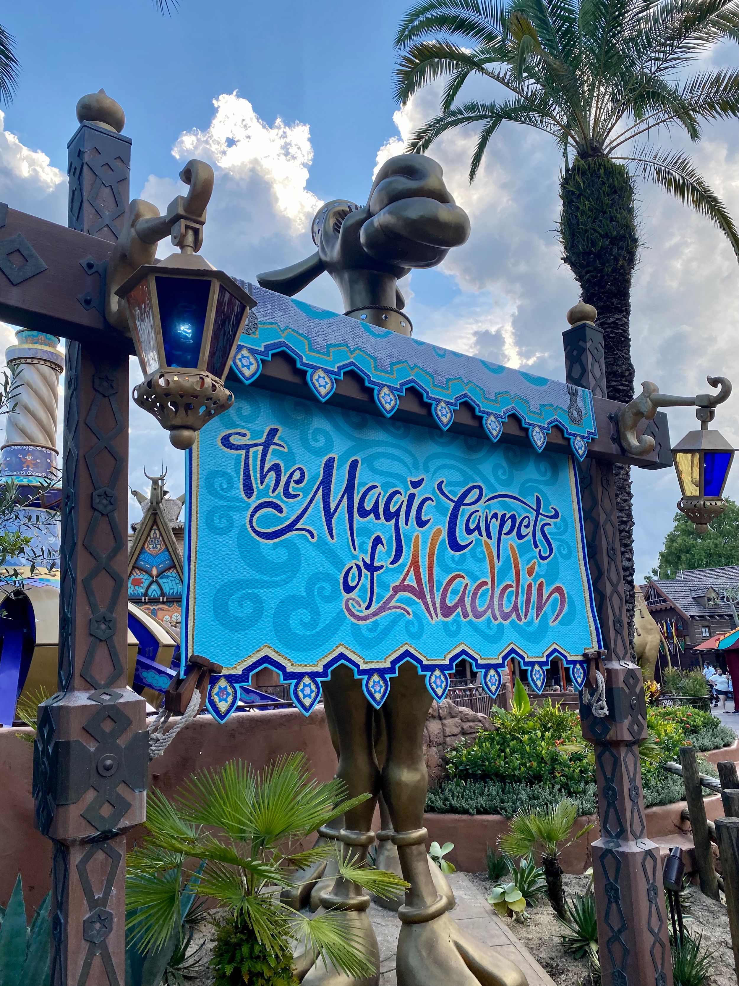 A blue sign for The Magic Carpets of Aladdin ride with decorative posts and palm trees at Disney theme park.
