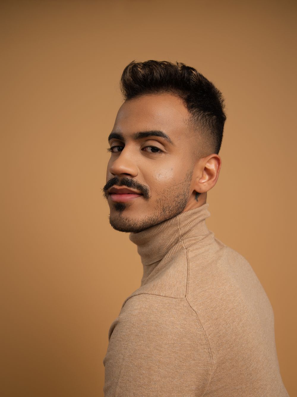Young man with mustache in beige turtleneck poses against warm brown background.