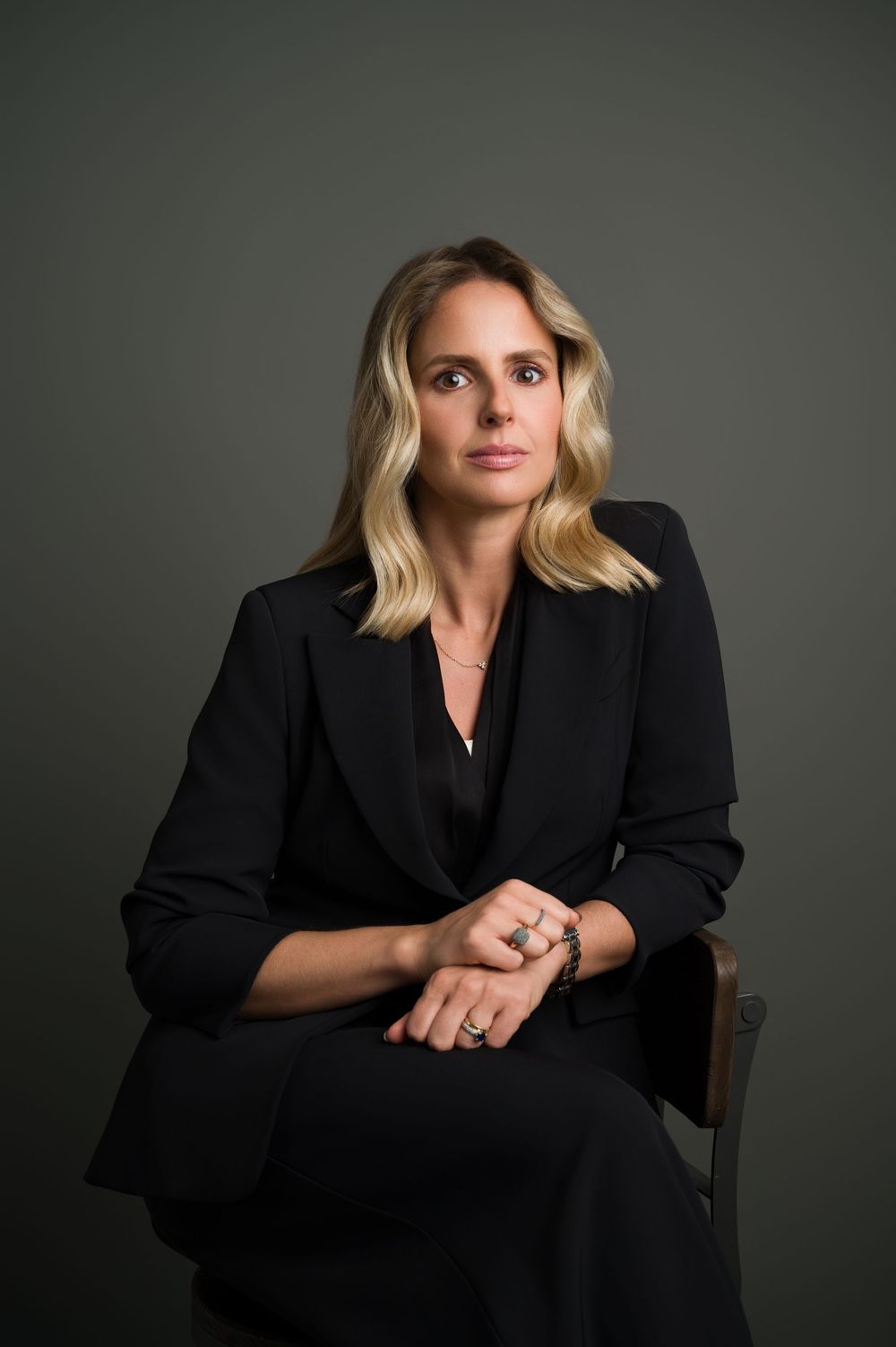 Professional woman in black blazer sitting with hands clasped, serious corporate portrait.