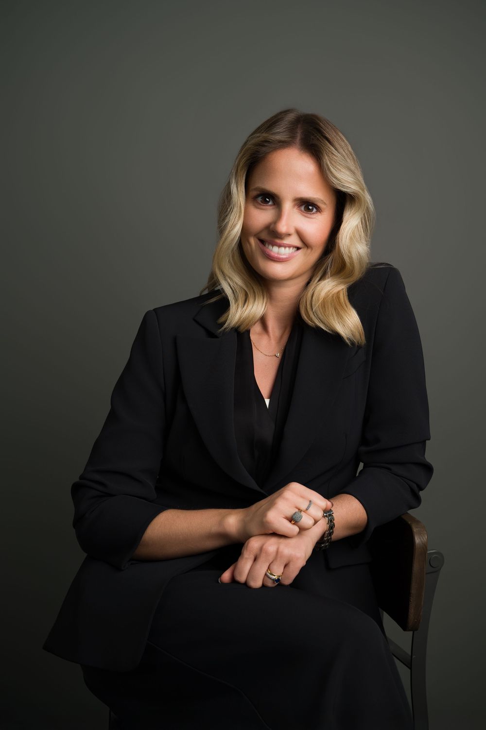 Smiling blonde woman in black business suit seated, professional headshot on dark background.