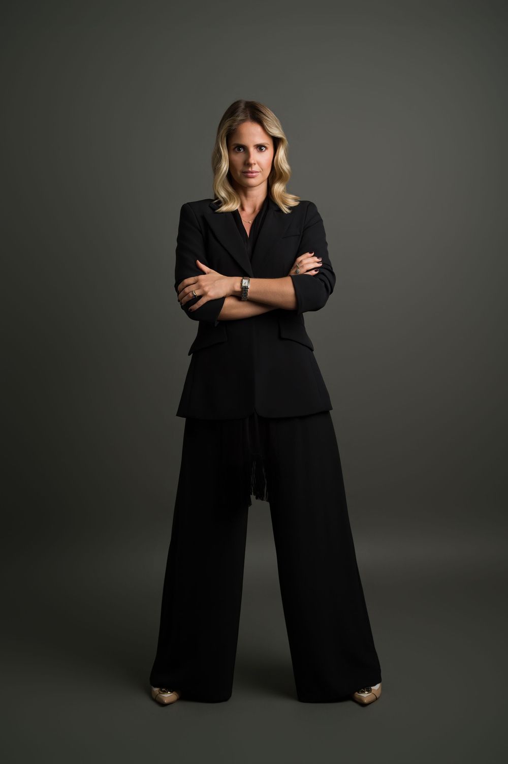 Blonde woman in black pantsuit standing with arms crossed, full-length professional portrait.
