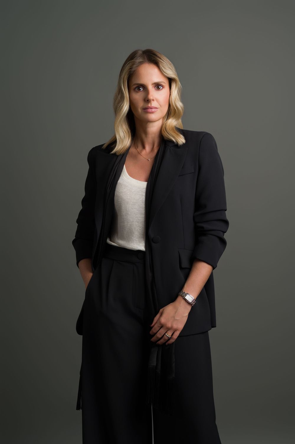 Blonde woman in black blazer and white top, hands in pockets, professional studio portrait.