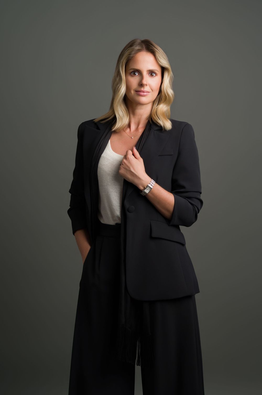 Blonde woman in black blazer holding lapel, confident professional studio fashion portrait.