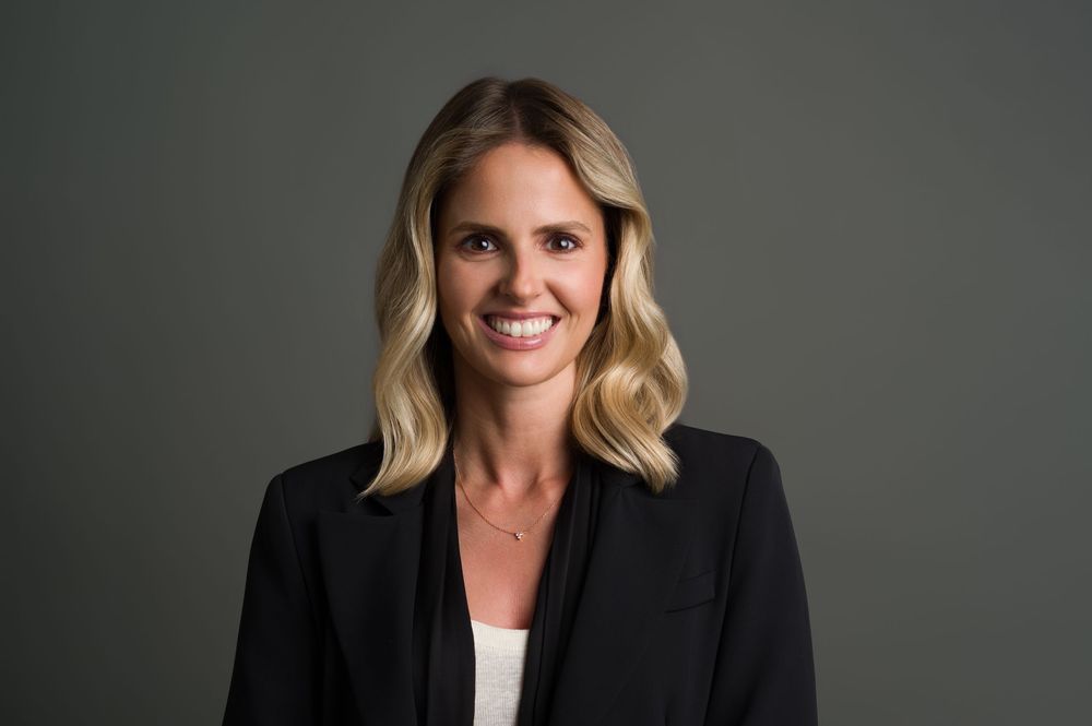 Professional headshot of a smiling blonde woman in a black blazer against a gray background.
