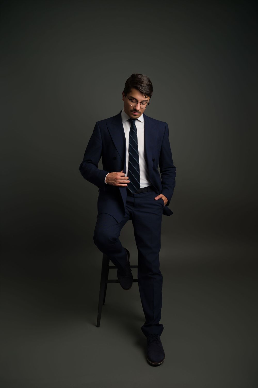 Man in navy suit and striped tie posing on a stool against a dark gray studio background.