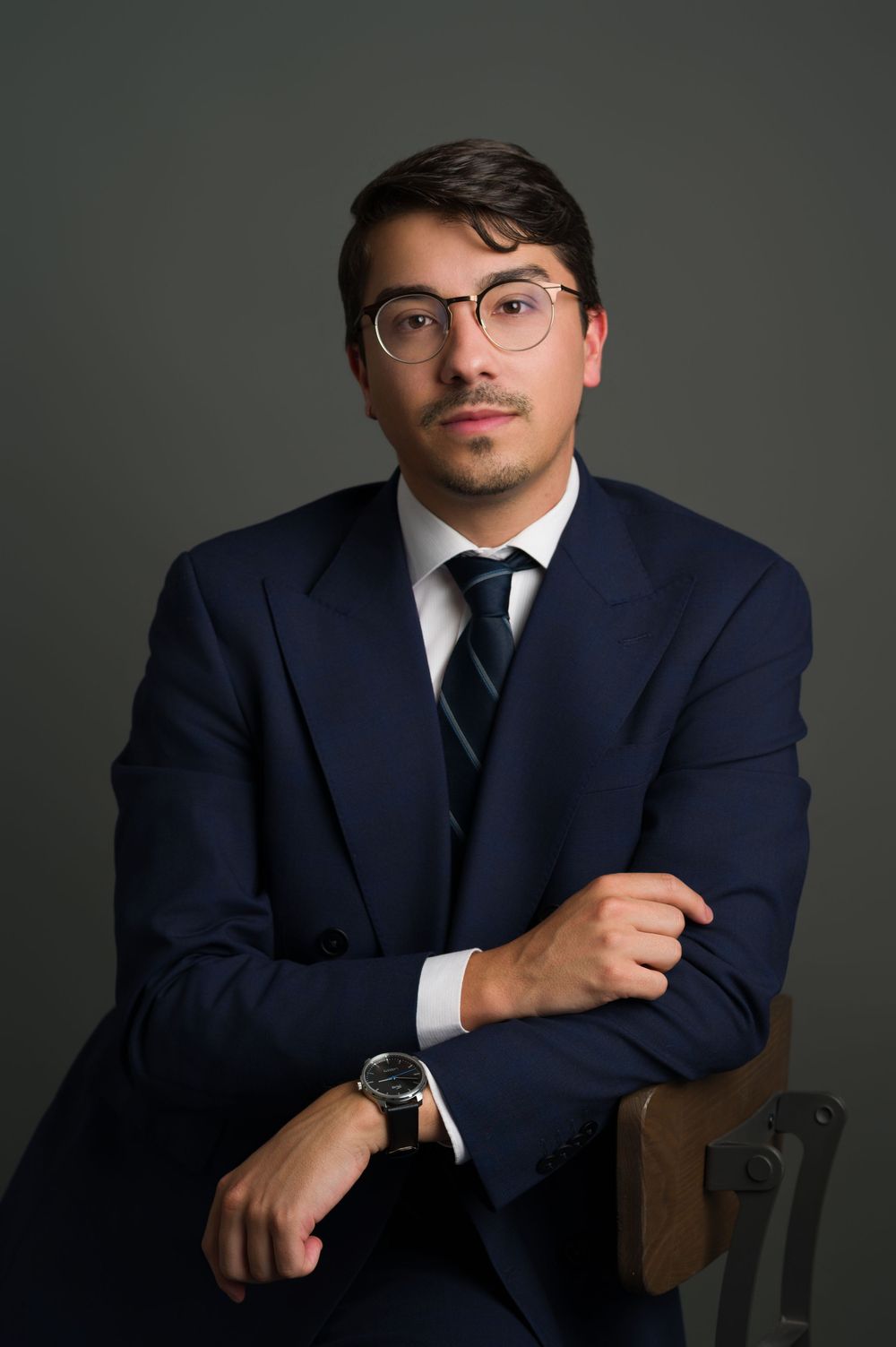 Professional portrait of a man in navy suit with crossed arms and watch, seated in a studio.