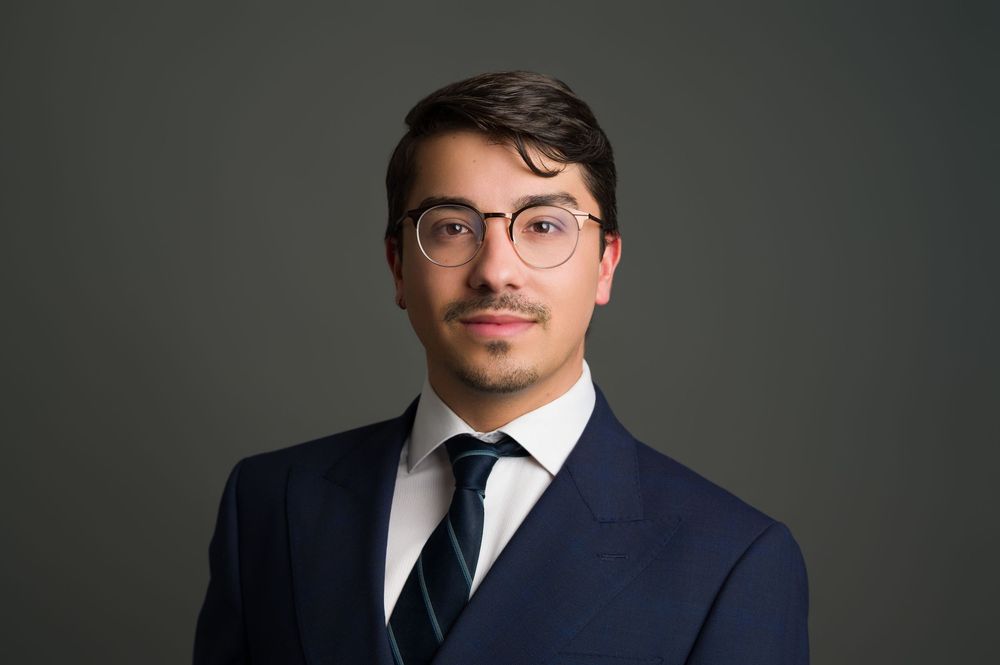 Close-up professional headshot of a young man in navy suit and striped tie wearing glasses.