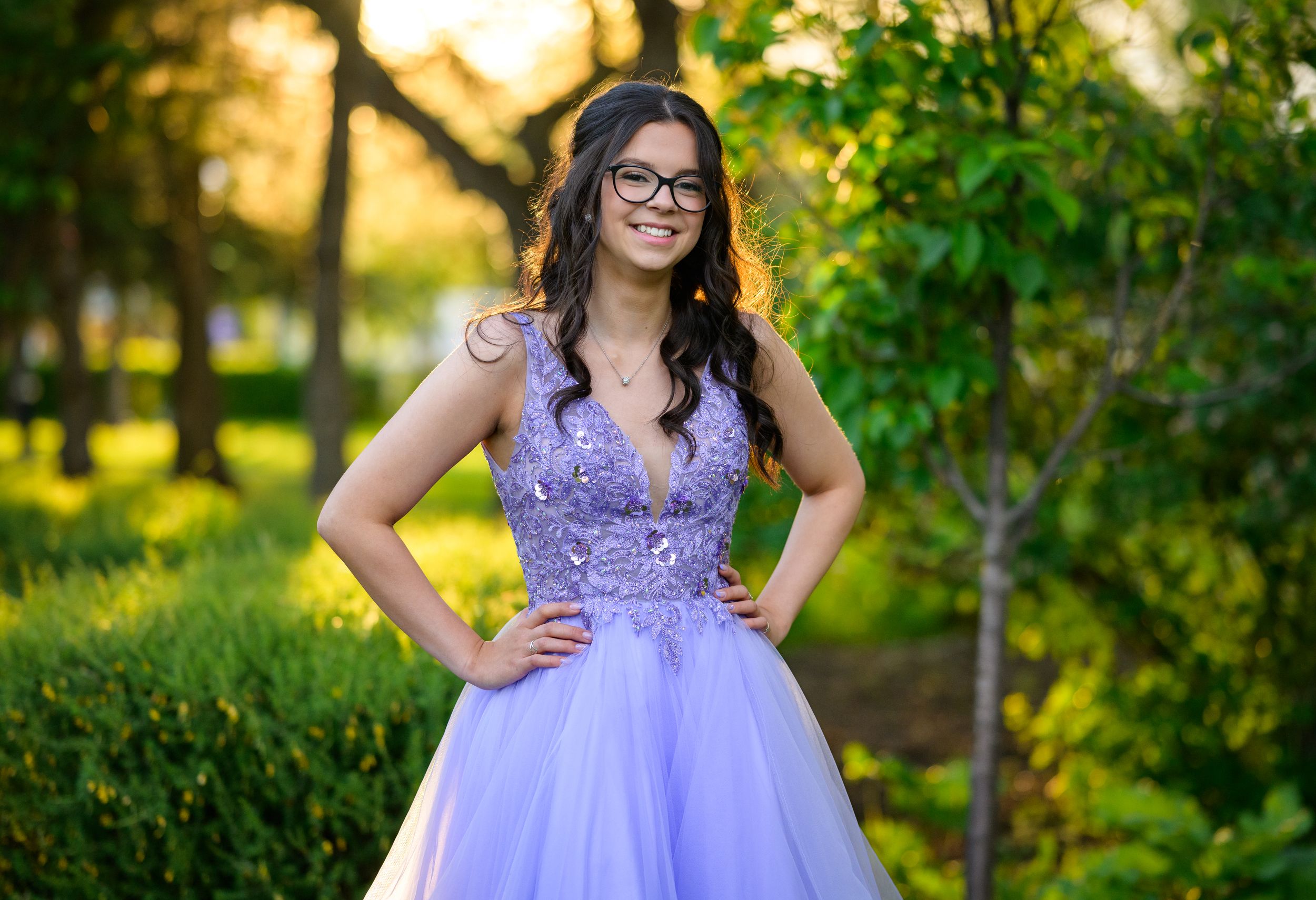A person in a lavender tulle prom dress poses outdoors in a garden setting during golden hour.